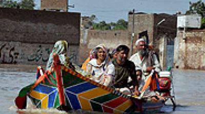 Four people ride a brightly painted wooden boat through a flooded urban area, with water covering the street and buildings visible in the background.