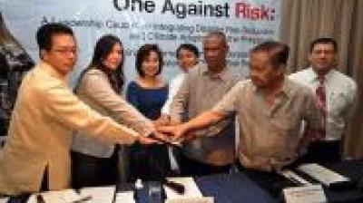 A group of six people stand together at a table, joining hands in the center, with a conference banner in the background.