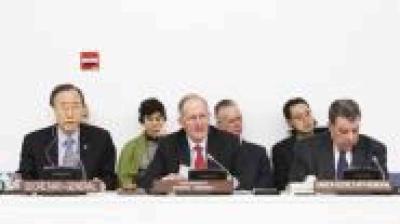 Six people sit at a conference table with microphones and nameplates in front of them, participating in a formal meeting.