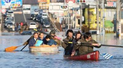 People paddle boats through a flooded city street as cars are partially submerged and traffic is halted in the background.