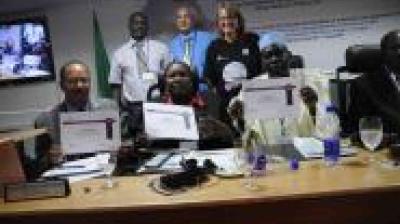 A group of people at a conference table hold certificates while others stand behind them, with documents and beverages on the table.