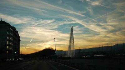 A city street at sunset with a tall water fountain in the distance and streaked clouds in the sky.