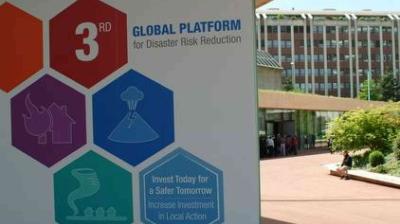 A sign for the 3rd Global Platform for Disaster Risk Reduction stands outdoors near a modern building, with icons illustrating various disaster types.