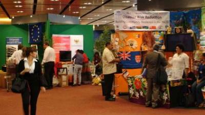 People interact at various booths during an indoor event focused on disaster risk reduction, with informational displays and banners visible.