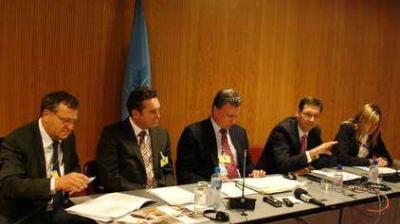 Five people in business attire sit at a conference table with documents, microphones, and water bottles in front of them, in a wood-paneled room with a blue flag in the background.