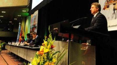 A man in a suit speaks at a podium on a stage, with several people seated at a long table beside him and flower arrangements in the foreground.