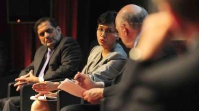 Three people in business attire sit on a panel, engaged in discussion, with one person gesturing while speaking and others listening attentively.