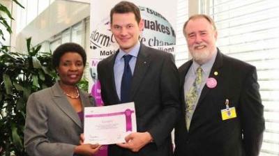 Three people in business attire pose indoors; one holds a certificate while the others stand beside him, smiling at the camera.