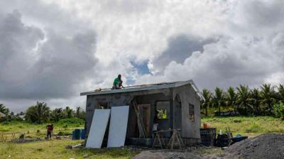 A person works on the roof of a small unfinished house in a grassy field, with tools and building materials scattered around and palm trees in the background under a cloudy sky.