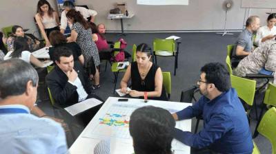A group of people sit around tables in a classroom, engaging in discussion and paperwork; maps and charts are spread out on the tables.