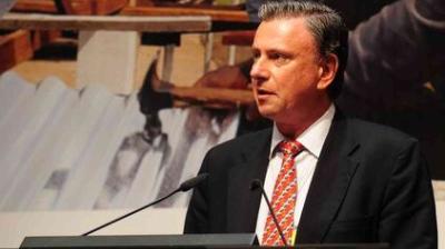 A man in a suit and patterned tie speaks at a podium with microphones, with a blurred background featuring construction materials.