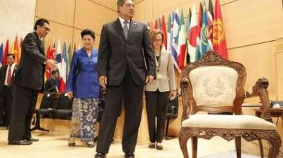 A group of formally dressed people walk past an ornate empty chair in a conference room with many international flags in the background.