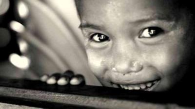 Close-up black and white photo of a smiling child with bright eyes, partially hiding behind a wooden surface.
