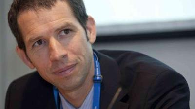 A man in a suit with a blue lanyard is looking slightly to the side and smiling during an indoor event or meeting.