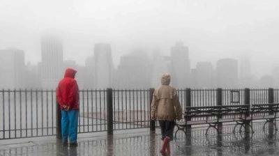 Two people in raincoats stand by a wet waterfront railing, with benches nearby and a city skyline obscured by heavy fog in the background.