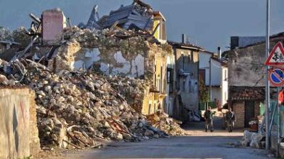 Collapsed building with rubble spilling onto a street after an earthquake; two people walk in the background near damaged structures.