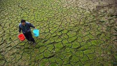 A person carrying two buckets walks across a cracked, dry, and moss-covered landscape, suggesting drought conditions.