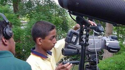 A young person adjusts a Panasonic video camera on a tripod outdoors, with another person nearby wearing headphones.