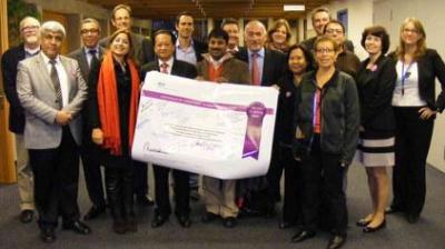 A group of people pose indoors, holding a large signed poster with text and graphics, while others stand around them, facing the camera.