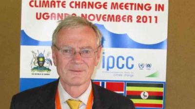 A man stands in front of a poster for a climate change meeting in Uganda, dated November 2011, with the IPCC logo and various flags displayed.