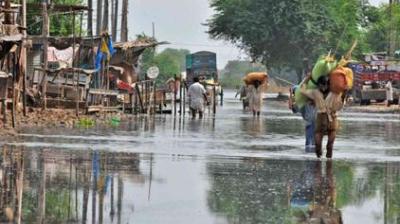 People walk through a flooded street lined with makeshift structures and trees, carrying belongings on their heads and shoulders.
