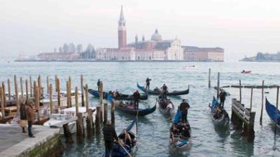 Gondolas with passengers navigate a canal in Venice, Italy, with the historic San Giorgio Maggiore island and church visible in the background.