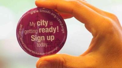 Hand holding a round purple button that reads, "My city is getting ready! Sign up today." and includes a website URL and campaign details.