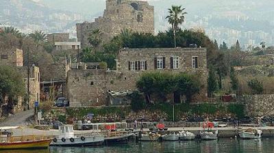 Several small boats are docked along a waterfront in front of stone buildings and historic ruins, with palm trees and a cityscape visible in the background.