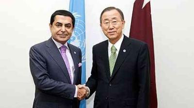 Two men in business attire shake hands in front of a United Nations flag and another flag in an official setting.