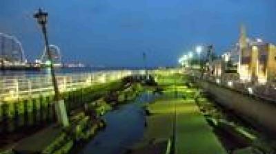 A seaside promenade at dusk with green-lit walkway, streetlights, buildings on the right, and a body of water on the left.