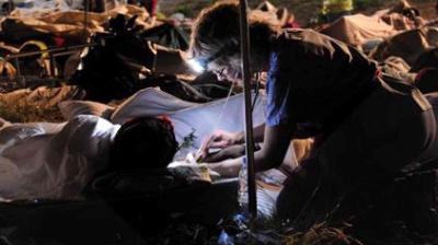 A person wearing a headlamp provides medical care to someone lying on a cot outdoors at night, surrounded by other patients and emergency supplies.