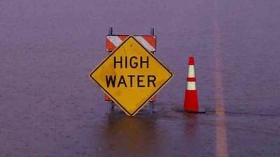 Yellow "HIGH WATER" warning sign and orange traffic cone partially submerged in floodwater.