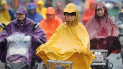 People wearing colorful rain ponchos ride bicycles and scooters in the rain, with water droplets visible in the air.