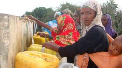 Women and children collect water from outdoor taps, filling yellow containers, with trees visible in the background.