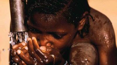 A child kneels and drinks water with cupped hands from a pipe, with droplets visible on their face and arms.