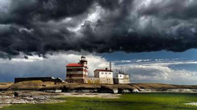 A lighthouse and adjacent buildings stand on a rocky shore under a dramatic, dark, and cloudy sky, with green water in the foreground.