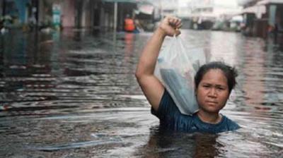 A woman wades through chest-deep floodwater on a city street, holding a plastic bag above her head to keep it dry.