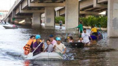 People ride boats through a flooded street under a bridge, navigating high water with oars. Others are seen wading or floating in the background.