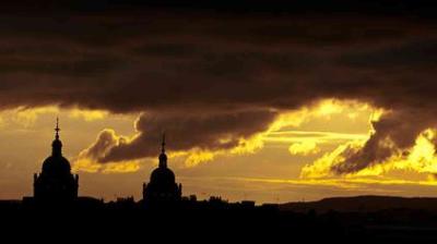 Dark clouds partially obscure a golden sunset sky, with the silhouette of two domed towers and city rooftops visible in the foreground.