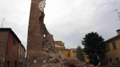 A partially collapsed brick clock tower stands amid rubble between buildings, showing significant earthquake damage under an overcast sky.