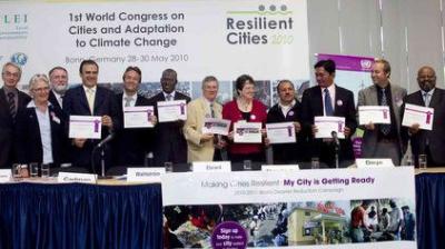 A group of people stand and sit at a table holding certificates at the 2010 Resilient Cities Congress in Bonn, Germany, with event banners displayed behind them.