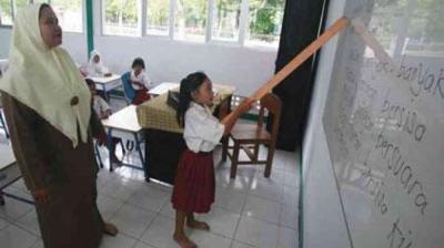 A girl in a school uniform points to words on a whiteboard with a stick while a teacher observes; other students are seated at desks in the classroom.