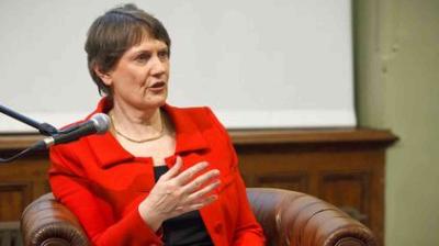 A person in a red jacket sits in a brown leather chair, speaking into a microphone with one hand raised, against a blurred indoor background.