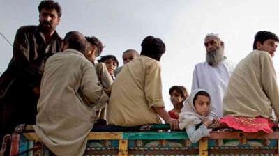 A group of men and children sit and stand on the back of a truck, with some looking toward the camera and others facing away, under a clear sky.