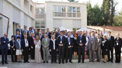 A large group of people in formal attire pose for a photo outside a building with a balcony and trees in the background.