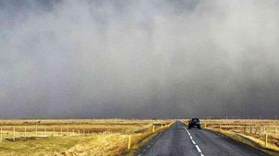 A car drives down a rural two-lane road with fields on either side under a sky filled with thick, gray fog or dust.