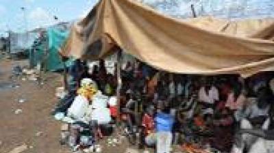 A group of people sit and stand under a large makeshift tent in a crowded outdoor area with scattered debris.