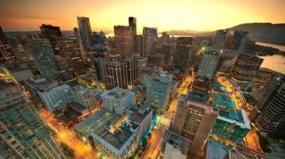 Aerial view of a city skyline at sunset, showing tall buildings, illuminated streets, and a body of water in the background.