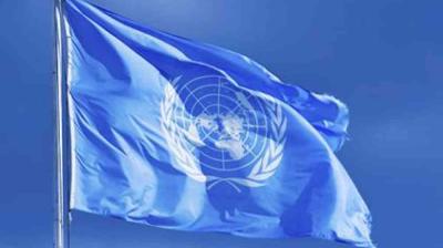 A United Nations flag featuring the UN emblem in white on a light blue background waves against a clear blue sky.