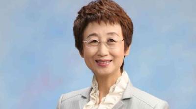 A woman with short brown hair, glasses, and a light gray blazer over a ruffled white blouse, smiling in front of a plain blue background.